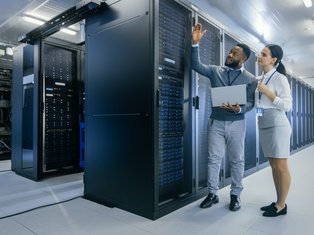 Two IT professionals in a data center discussing server infrastructure, with one holding a laptop and pointing at a server rack