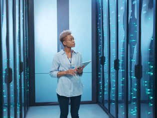 A female IT professional holding a tablet stands in a server room surrounded by illuminated data servers with glowing blue lights