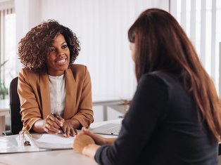 Two professional women engaged in a business meeting in a modern office space, one smiling and holding a pen while discussing documents on the table