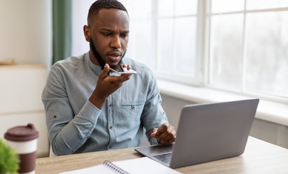 A young man sitting at a desk with a laptop, speaking into his smartphone with a focused expression, while a notebook and coffee cup are nearby